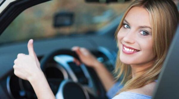 Woman driving car with thumbs-up, smiling confidently.